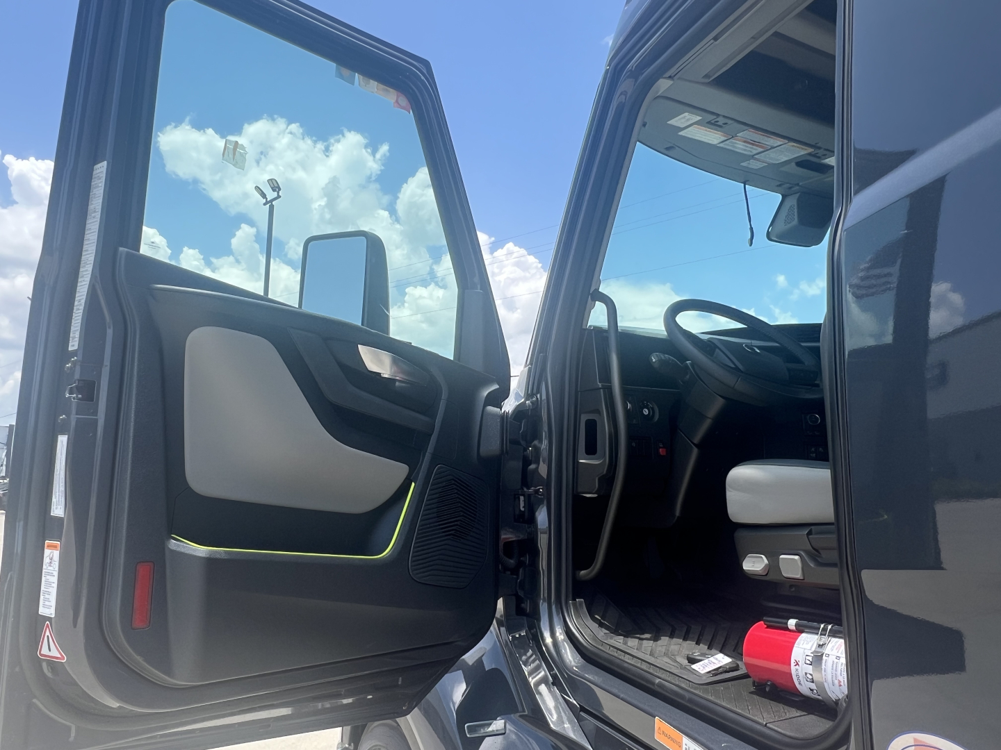 Volvo VNL64T660 cab interior with open driver door showing steering wheel and dashboard; exterior mirror and blue sky reflected; red fire extinguisher visible near door.