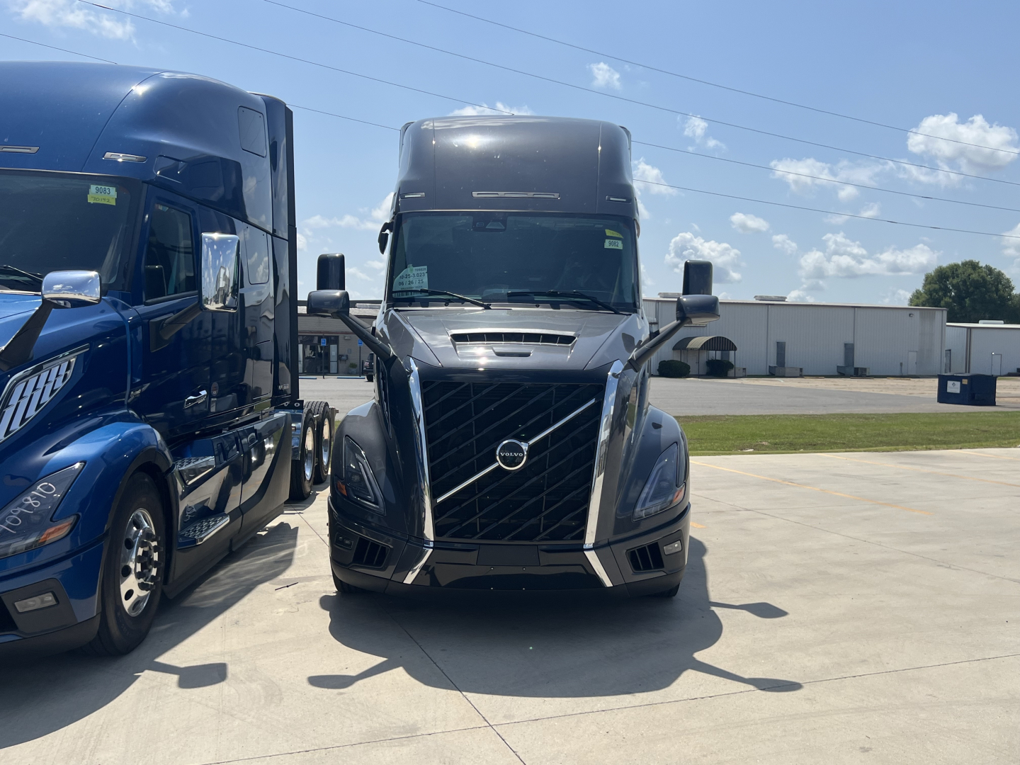Front view of a 2026 Volvo VNL64T660 truck in a lot, flanked by another blue truck on the left; visible Volvo emblem on grille; clear sky.