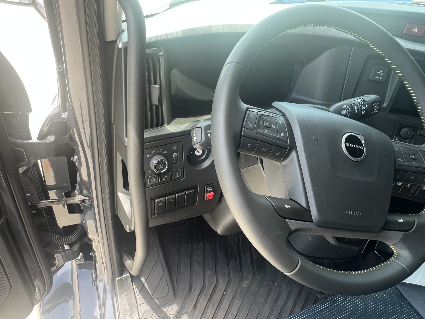 Volvo truck interior showing steering wheel with Volvo logo, left dashboard controls, and entry area; visible panels and pedals.