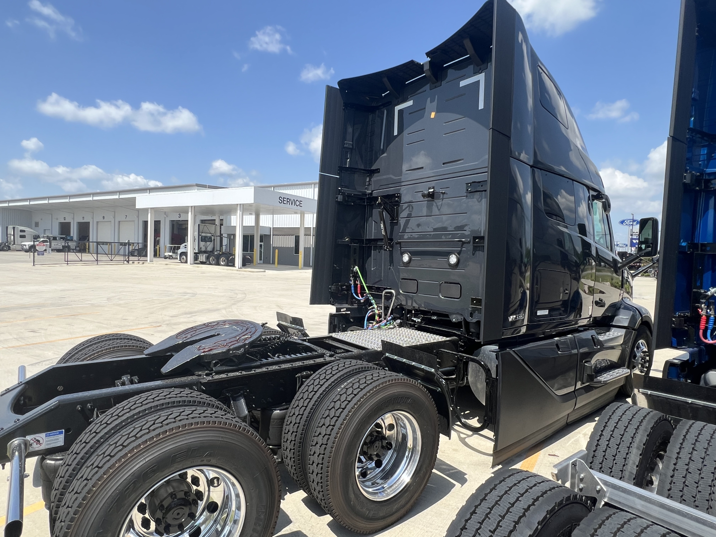 Volvo VNL truck cab and chassis in a lot, rear view showing dual rear axles and fifth wheel area, black exterior, blue equipment on right, clear sky