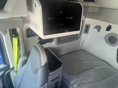 Interior of a new Volvo VNL sleeper cab showing a mounted overhead compartment and a bed area with a gray mattress, seat in foreground, and control panels on the wall.