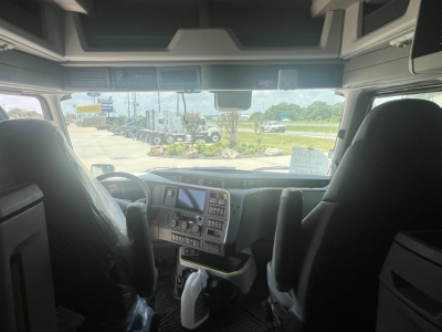 Cab interior of a Volvo truck with steering wheel, dashboard, and seats; windshield shows outdoor lot with other trucks and trailers.