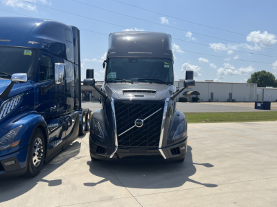 Front view of a 2026 Volvo VNL64T660 truck in a lot, flanked by another blue truck on the left; visible Volvo emblem on grille; clear sky.