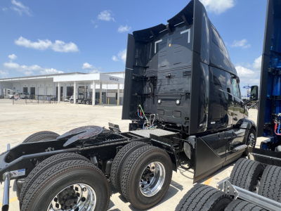 Volvo VNL truck cab and chassis in a lot, rear view showing dual rear axles and fifth wheel area, black exterior, blue equipment on right, clear sky