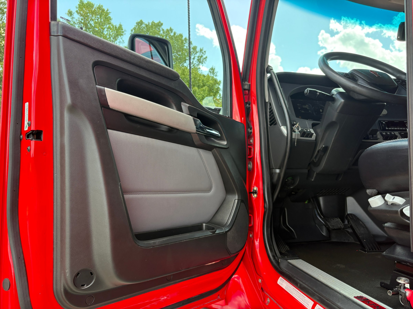 Truck cab interior door panel and steering wheel area of a red 2022 Kenworth T680, open driver door showing dashboard and floor space.