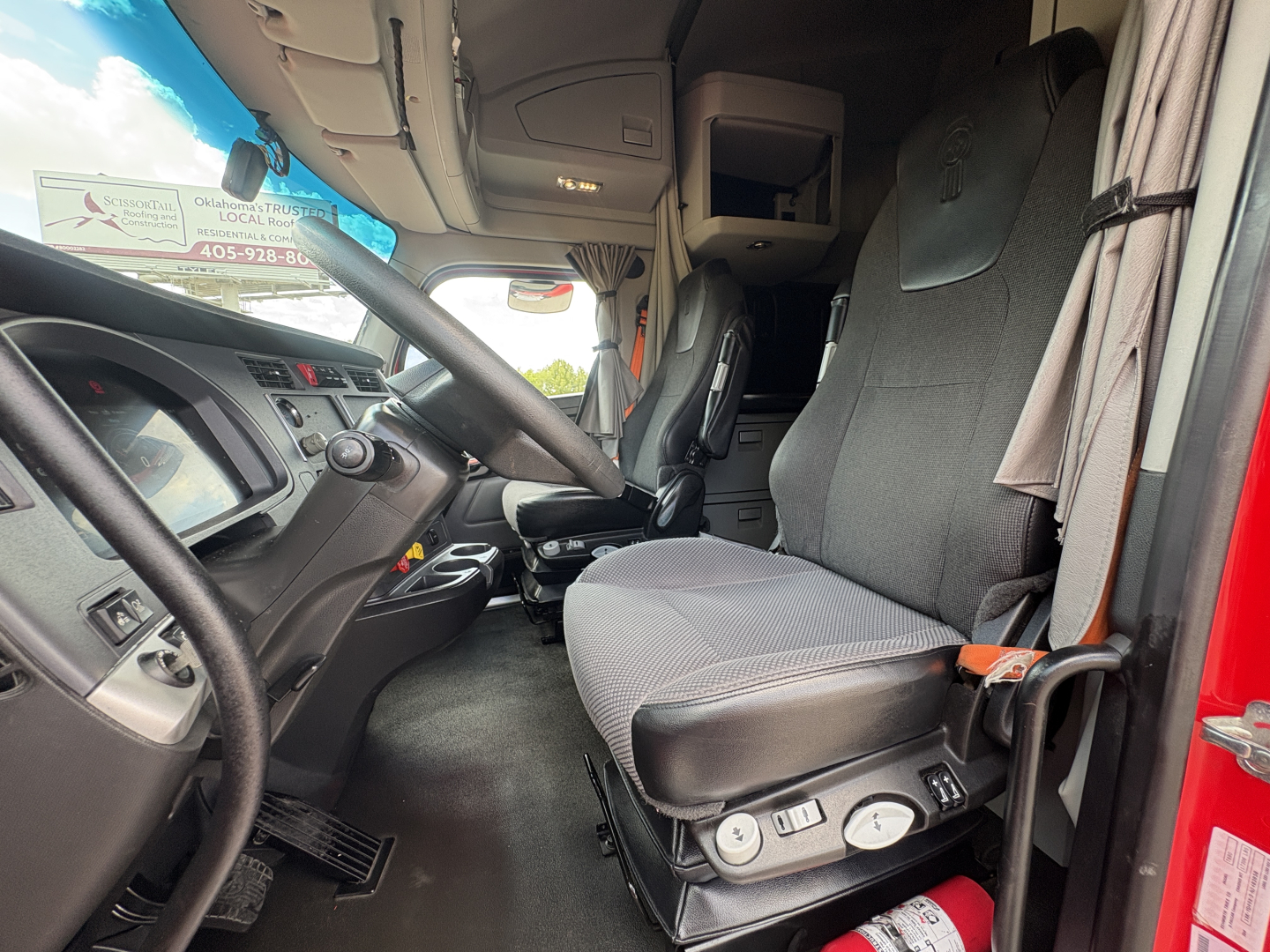 Cab interior of a 2022 Kenworth T680 showing driver's seat with controls, steering wheel, and dashboard visible in daylight.