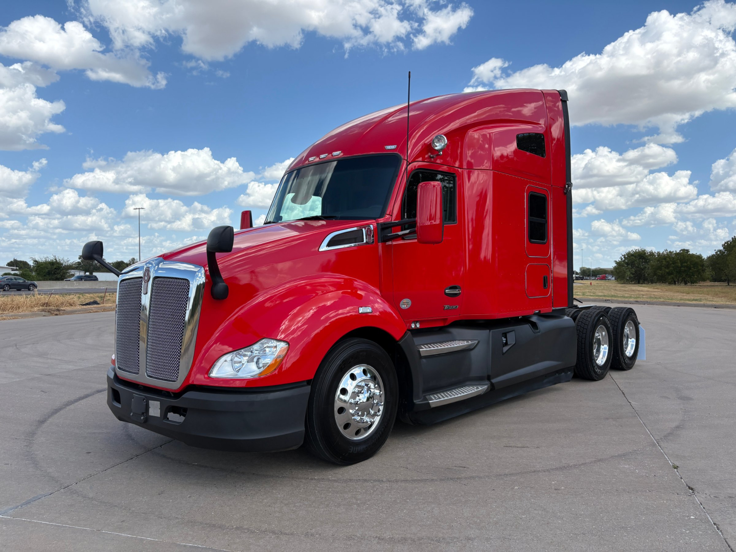 Kenworth T680 tractor unit in red, side view showing sleeper cab, front chrome grille, dual rear axles, black bumper, silver wheels, blue sky with clouds.