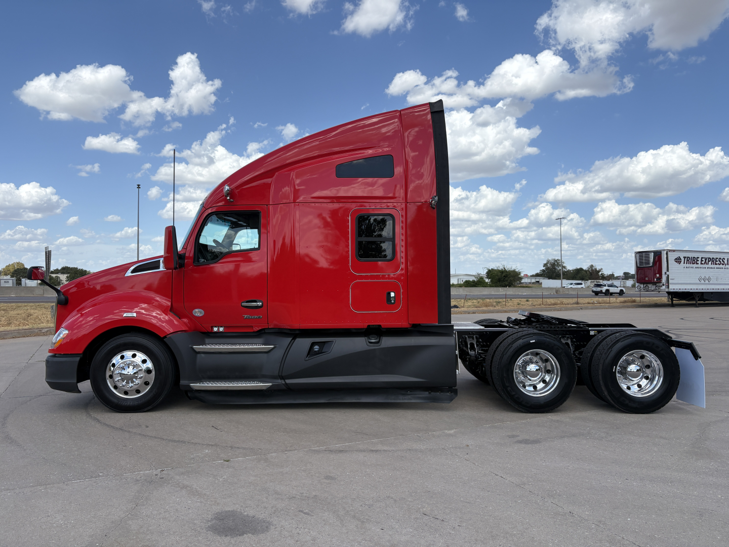 Kenworth T680 semi-truck cab in red side view showing sleeper, fuel tanks, and triple rear axles.