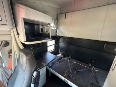 Kenworth T680 sleeper cab interior showing bunk area with seat, storage cabinet, and seatbelt anchor, label panels, and small window.