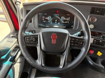 Truck cab steering wheel and dashboard view showing Kenworth logo on steering wheel and digital instrument cluster.