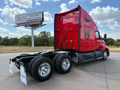 Kenworth T680 tractor in red, side view showing rear wheels and cab. No visible text on the truck, but a roadside sign in background reads Oklahoma’s TRUSTED... with phone number 405-928-8089.
