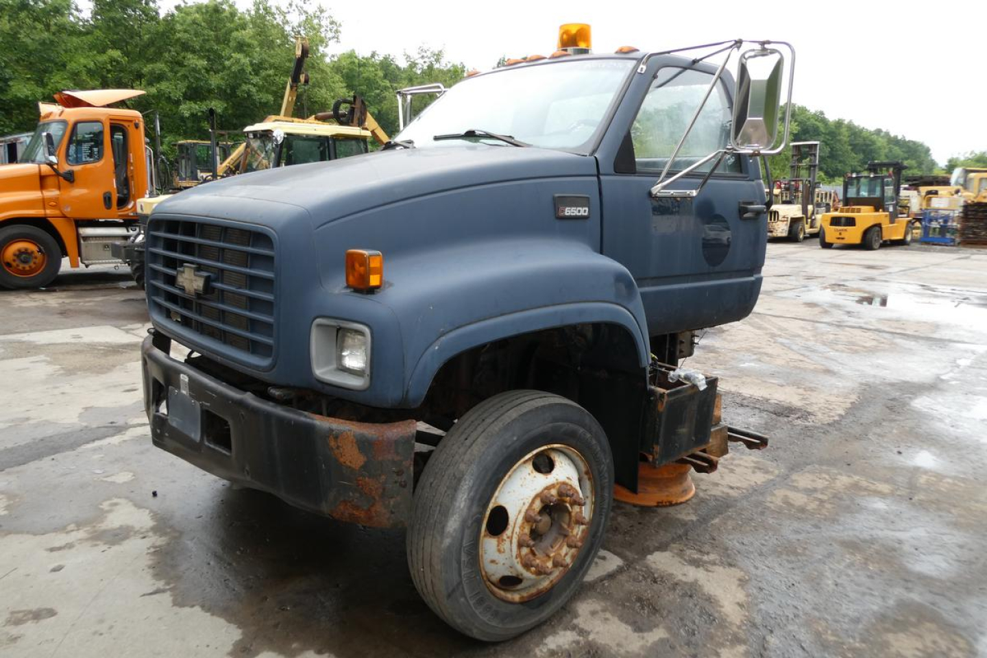 Chevrolet C6500 cab and chassis truck from 2001 with blue body, open front passenger door, and rusted front bumper; single front wheel visible with hub and exposed suspension.