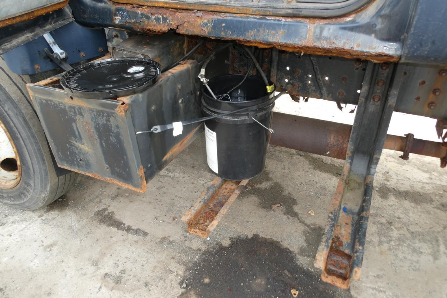Truck chassis area showing a metal box and cylindrical object mounted under the cab near the left side, with exposed frame rails and a rusted support member.
