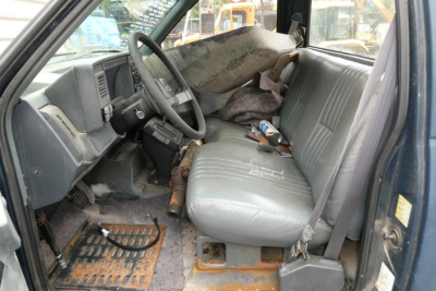 Cab and chassis interior showing steering wheel, bench seat, and worn dashboard of a 2001 Chevrolet C6500. Textured gray seats with seat belt, debris on floor and dirty floor mat.