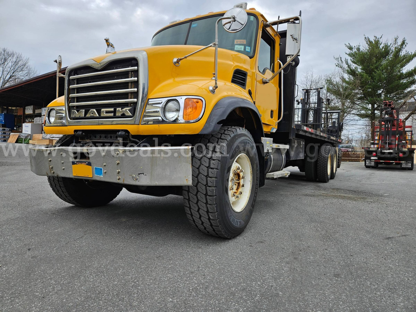 Mack CV713 Granite truck in yellow with flatbed, front view showing grille and bumper, large tires, and side mirrors.