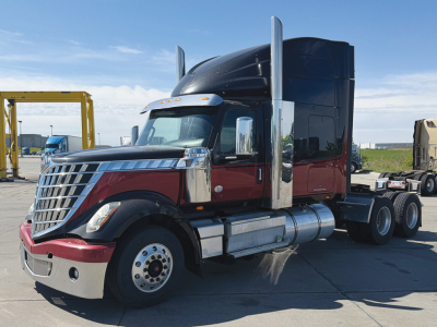 2021 International LoneStar truck with maroon lower body and black sleeper cab, chrome exhaust stacks, large front grille, side fuel tanks; truck in a dealership yard.