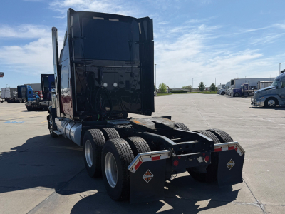 Truck chassis in yard view showing rear of tractor with fifth-wheel and tandem axles. Visible diamond placards and mudflaps with warning labels.