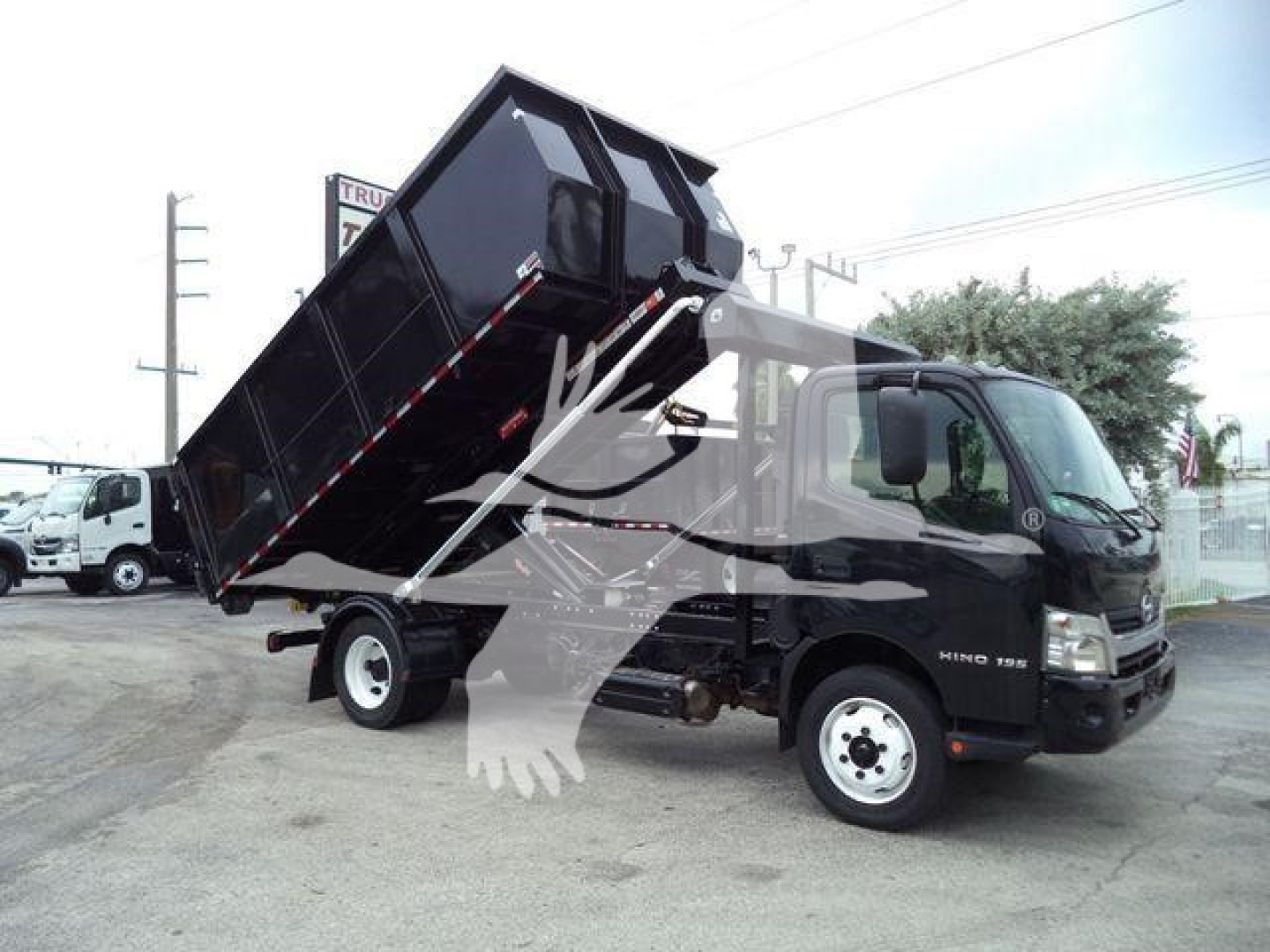 2019 Hino 195 dump truck with tilted black dump bed and white frame, cab side shows Hino badge. Vehicle in lot with other trucks in background.