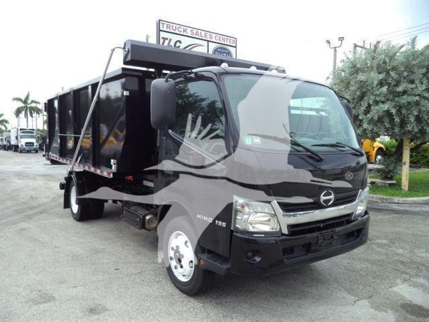 2019 Hino 195 roll-off truck in a used condition, front cab view with black dump body, Hino emblem on grille, side mirrors, and load bed visible.