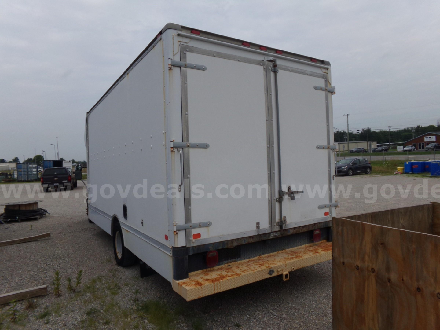 Box truck rear view showing white cargo box with double rear doors, hinges and latches, rust on bumper plate, gravel lot in background.