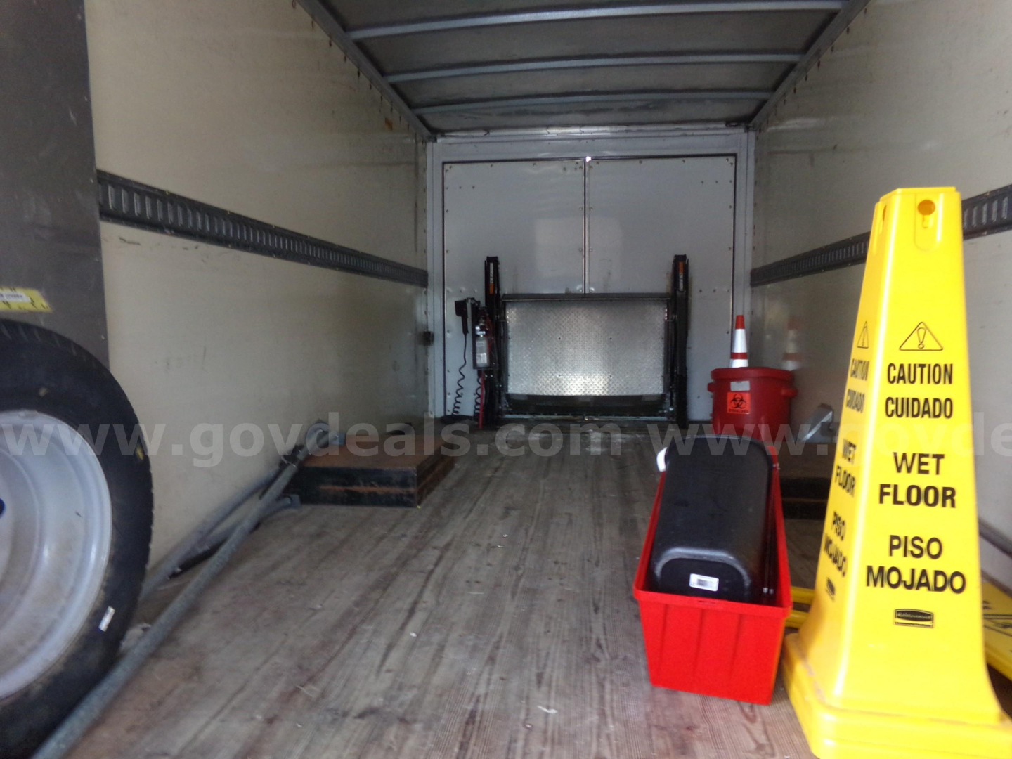 Truck bed interior with loading equipment and yellow caution sign, wheel on left, red toolbox, wet floor sign, and white rear door.