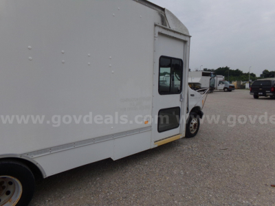 Box truck side view showing white cargo body and cab with window, with gravel lot and other trucks in background.
