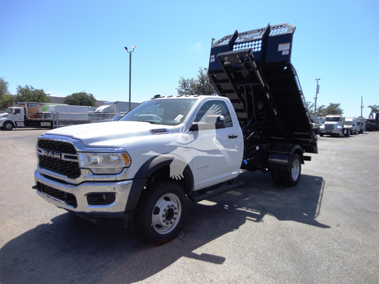 Ram 5500 truck with a raised dump bed in a dealership lot, side view showing white cab and black hooklift bed.