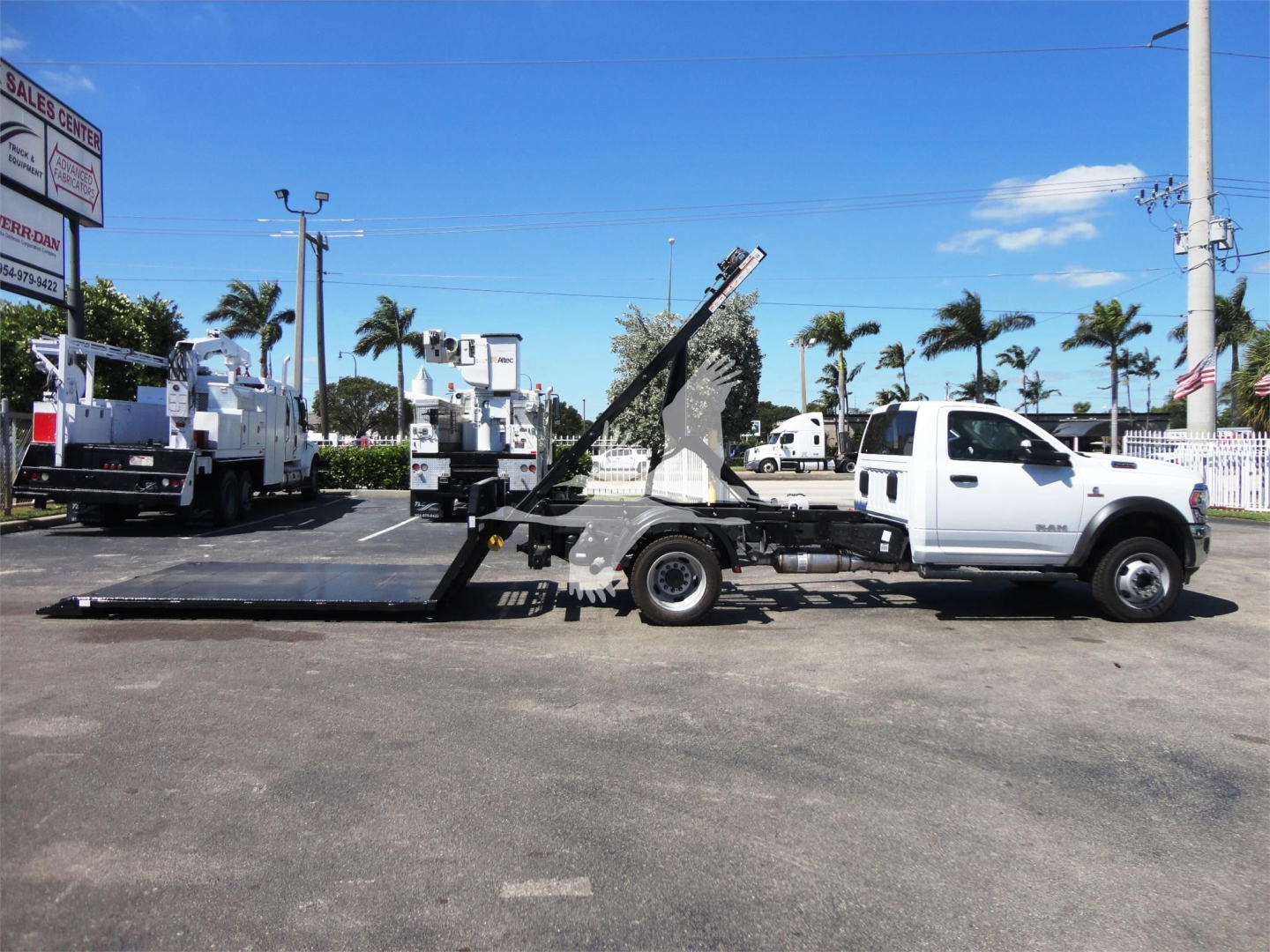 Ram 5500 HD tow truck with long flatbed and hydraulic tilt bed in a dealership lot, white cab, black equipment, palm trees in background.