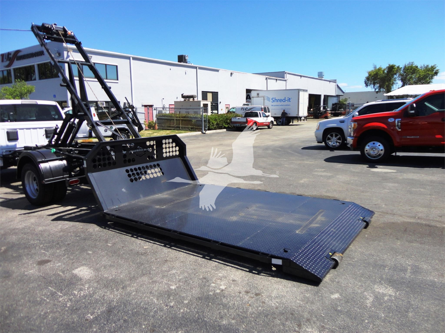 Ram 5500 heavy-duty hooklift truck with a flatbed ramp in a dealer lot, other trucks in background.