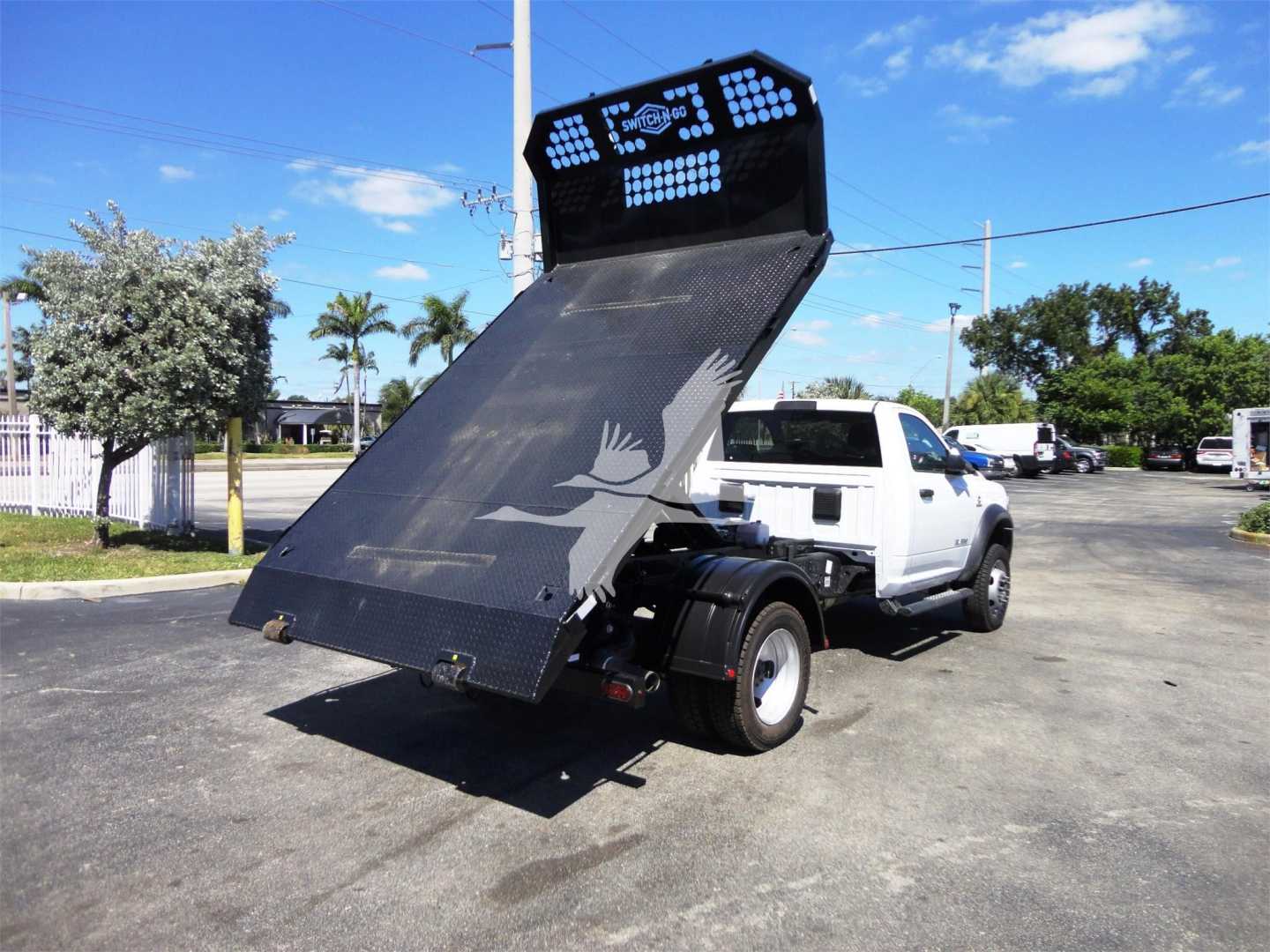 Ram 5500 dump bed truck with elevated hydraulic tilt bed, hooklift configuration, white cab, rear view showing black steel bed and tailgate plate.