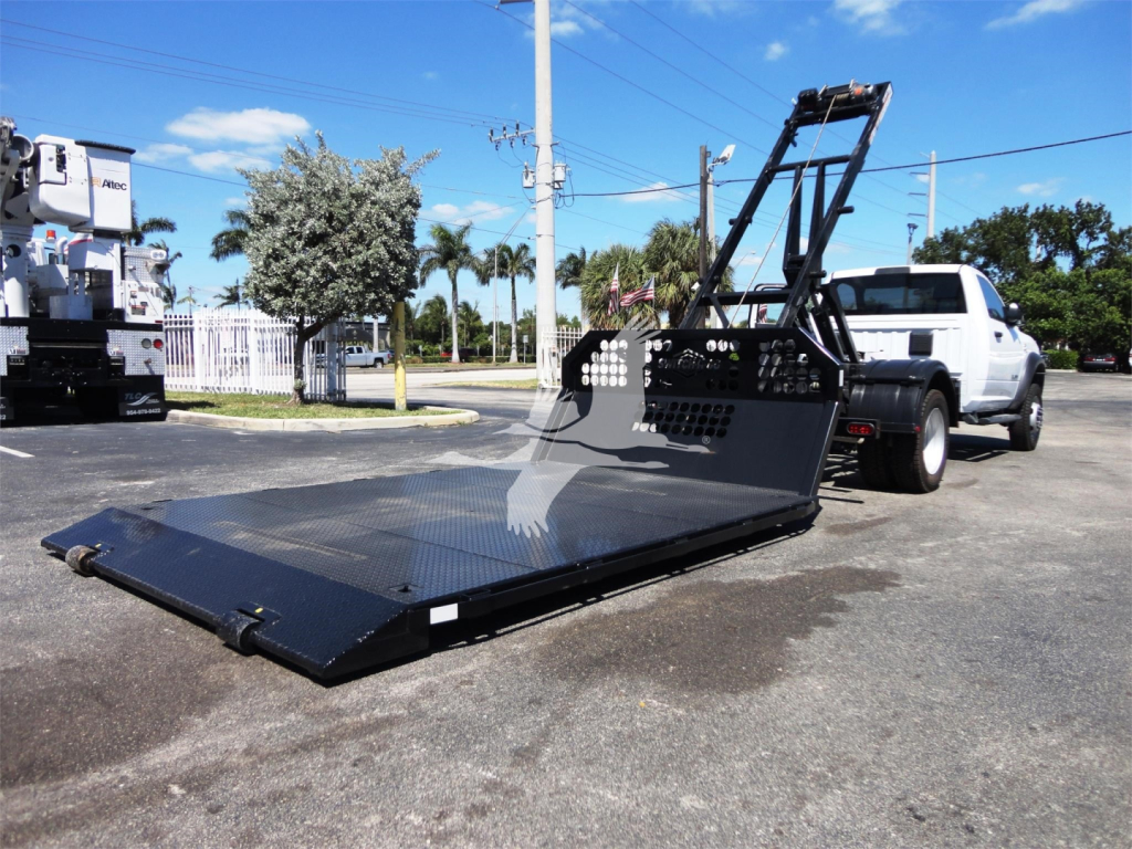 Ram 5500 HD truck with a large flatbed hooklift platform extended, rear ramps folded up, white cab, in a dealership lot.
