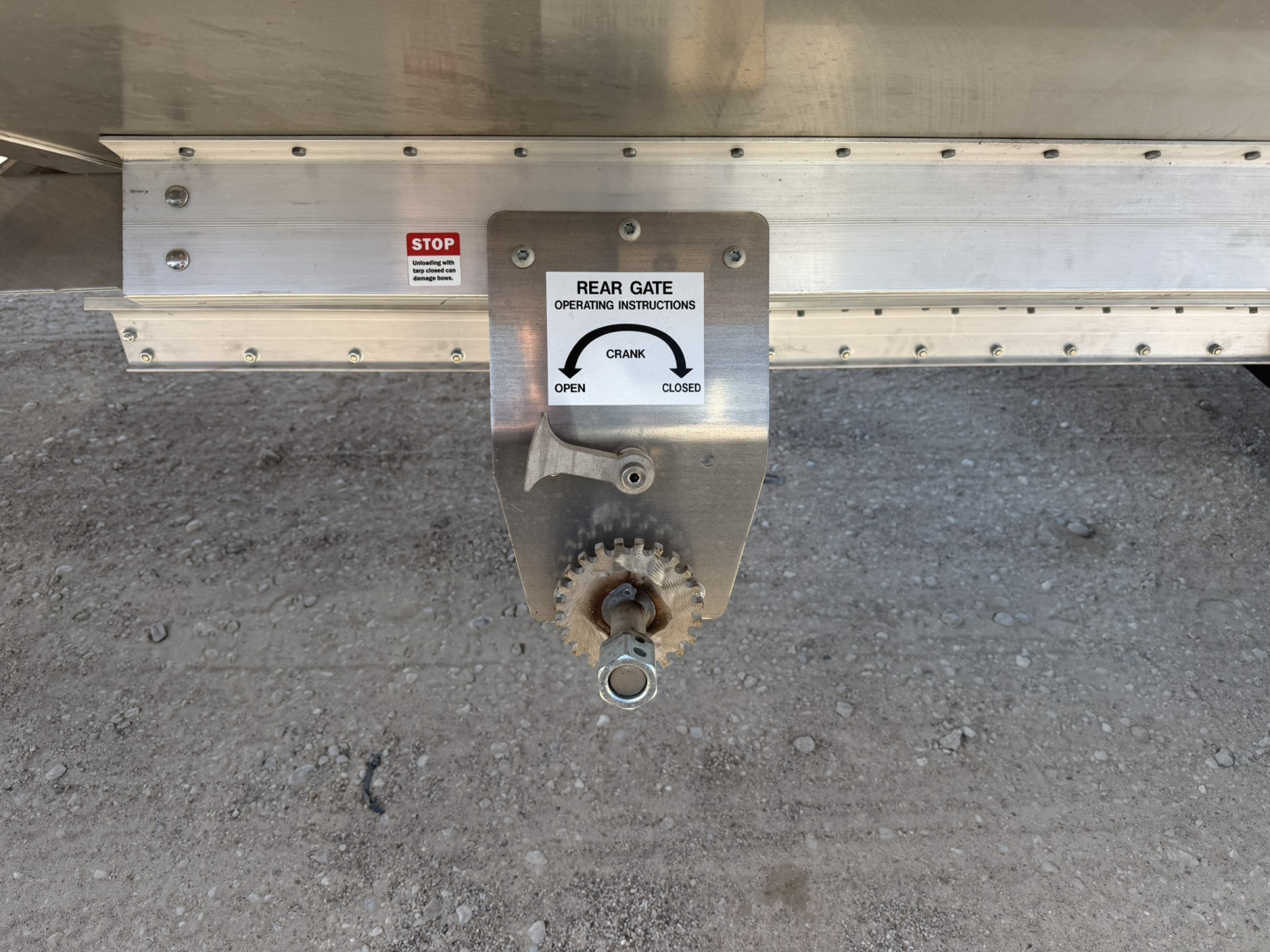 Trailer hitch and rear gate crank box on bottom of 40' aluminum grain hopper trailer; visible label reads REAR GATE OPERATING INSTRUCTIONS with CRANK OPEN CLOSE and hand wheel.