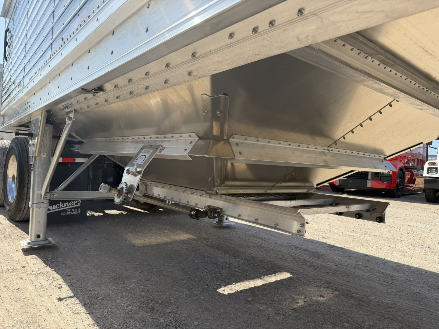 Trailer undercarriage showing aluminum grain hopper with corrugated sidewalls and landing gear. Visible text on plate reads 'Stoughton' and metal brackets with rivets.