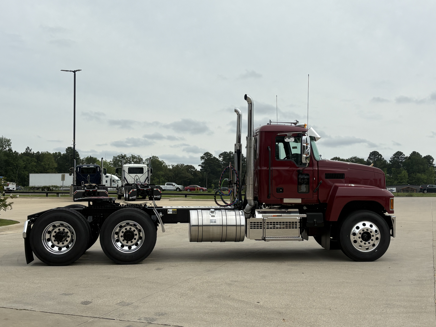 Truck chassis of a 2027 Mack PI64T with maroon cab and dual rear axles, side view showing fuel tanks and exhaust stacks.