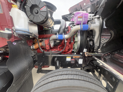 Truck engine bay showing diesel engine with red block, silver exhaust, coolant reservoir, purple coolant/DEF reservoir, hoses, and radiator components. 