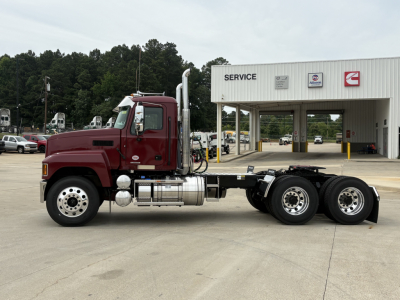 Truck chassis with a red Mack PI64T daycab, side view showing dual rear axles, fuel tanks, exhaust stack, and service bay building in background.