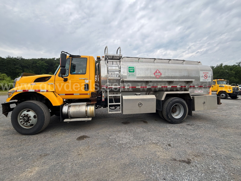 Tank truck with yellow cab and silver cylindrical tank showing warning labels and access steps on the side.