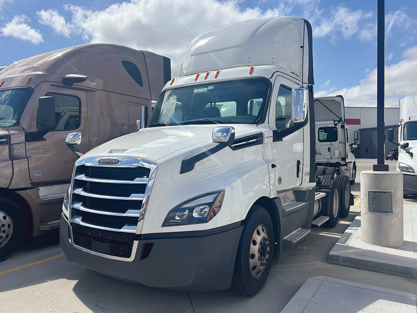 Truck cab with white Freightliner Cascadia 126 in a dealership lot, front grille visible, side mirrors, and other trucks in background.