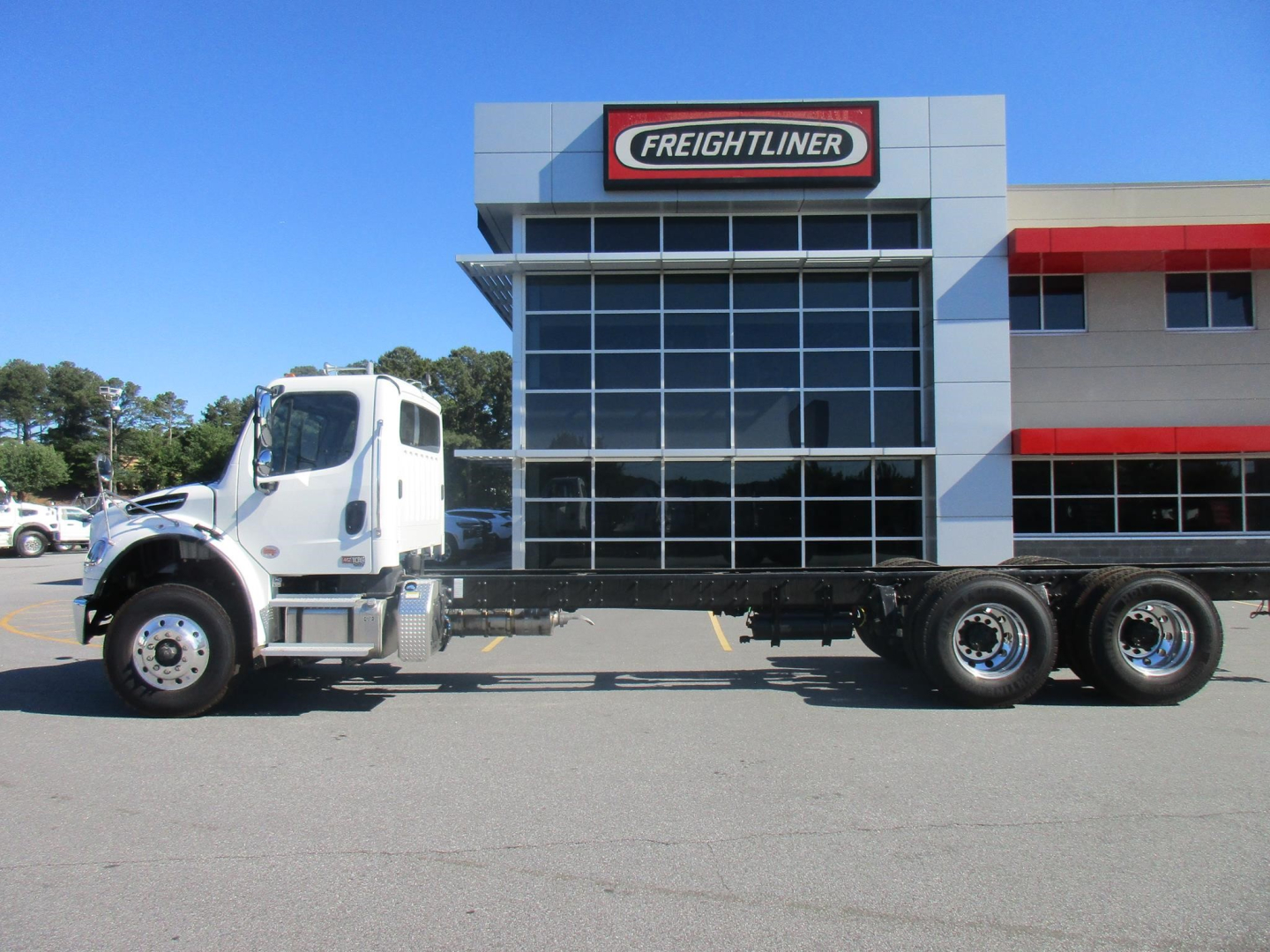 Freightliner Business Class M2 106 Plus cab and chassis truck side profile in dealership lot with large glass showroom behind.