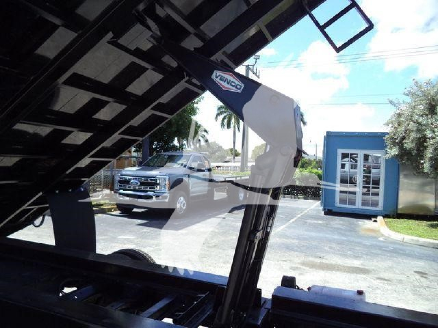 Ford E-450 dump truck in-service with hydraulic dump bed and label 'VANCO' on hydraulic arm, rear of cab visible, background shows pickup and building.