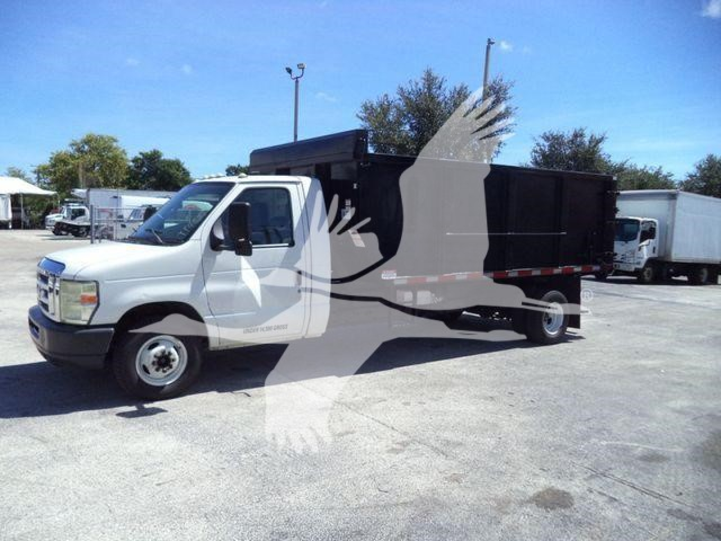 Dump truck body on a white Ford E-450 cab; black dump bed with hydraulic system visible; clear weather, other trucks in background.