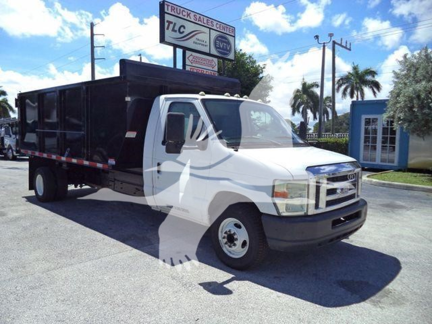 Ford E-450 dump truck side view showing white cab and black dump body with TLC sign in background.