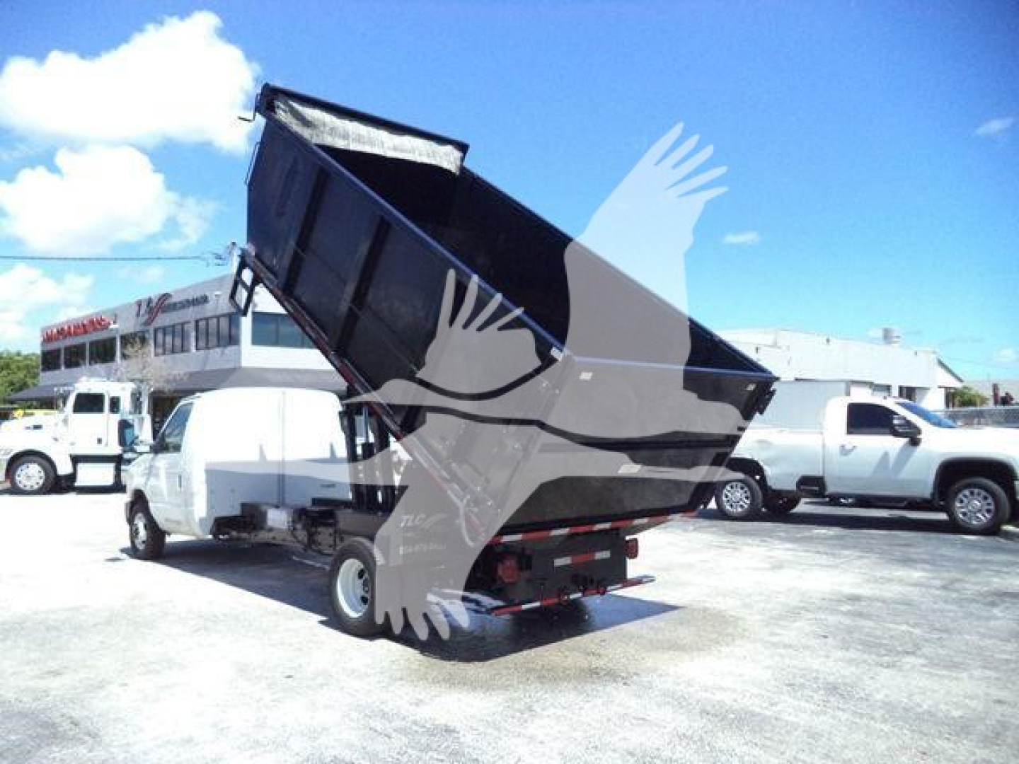 Ford E-450 dump truck with a raised black dump bed in a parking lot. Visible text: none readable on the truck body; background shows other trucks.