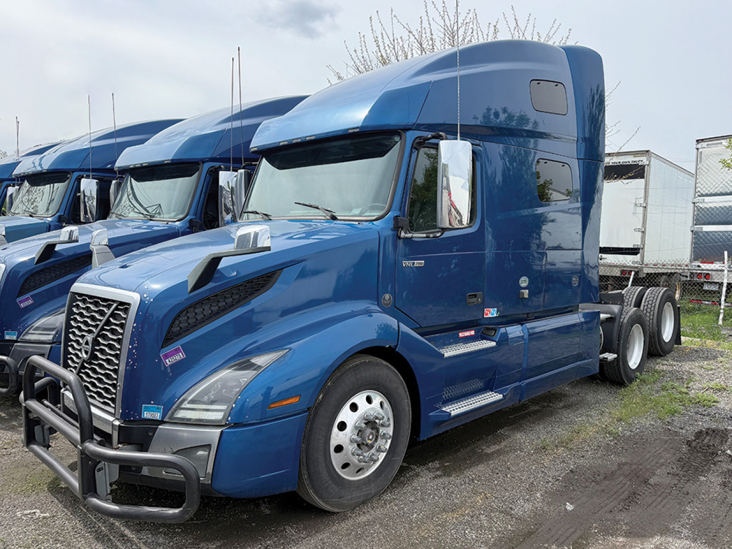 Blue Volvo VNL 760 tractor in yard with sleeper cab, side view showing front grille and bumper, no text clearly visible besides small labels on door and hood.
