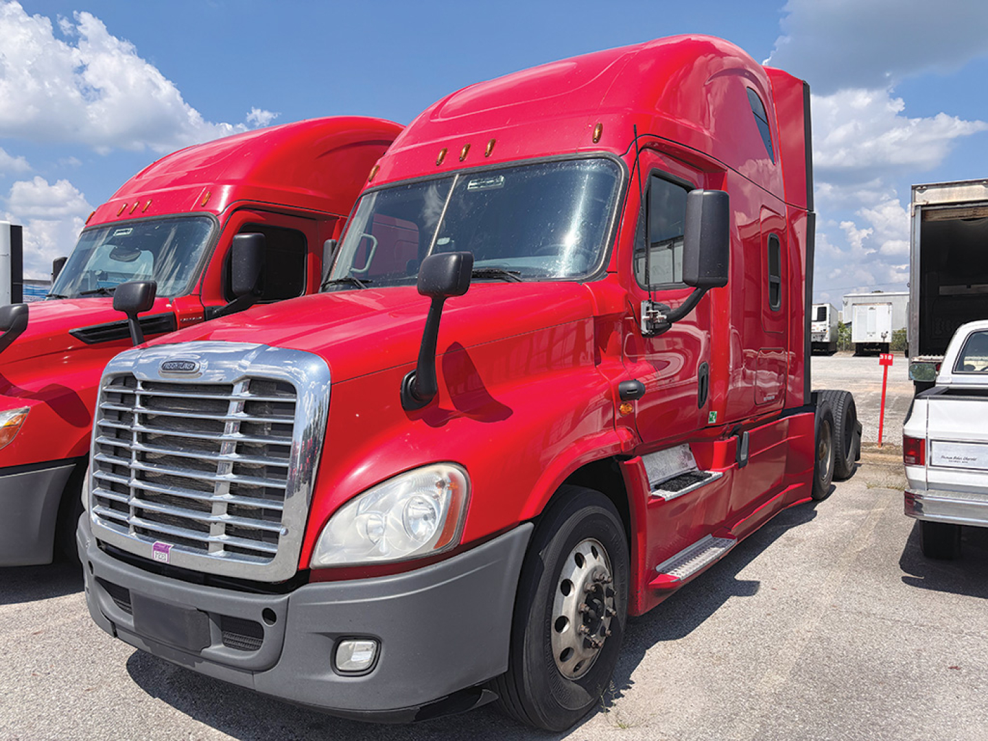 Freightliner Cascadia125 semi-truck in red foreground, cabin with large grille, black mirrors, side steps, and wheel exposed; other red trucks visible in background.