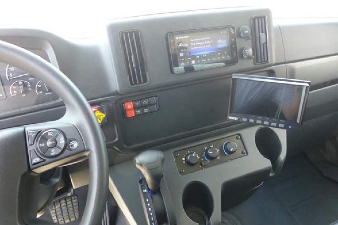 Cab interior of a Freightliner M2 106 Plus showing steering wheel, dashboard with touchscreen infotainment, adjustable monitor, gear shifter, and cup holders.