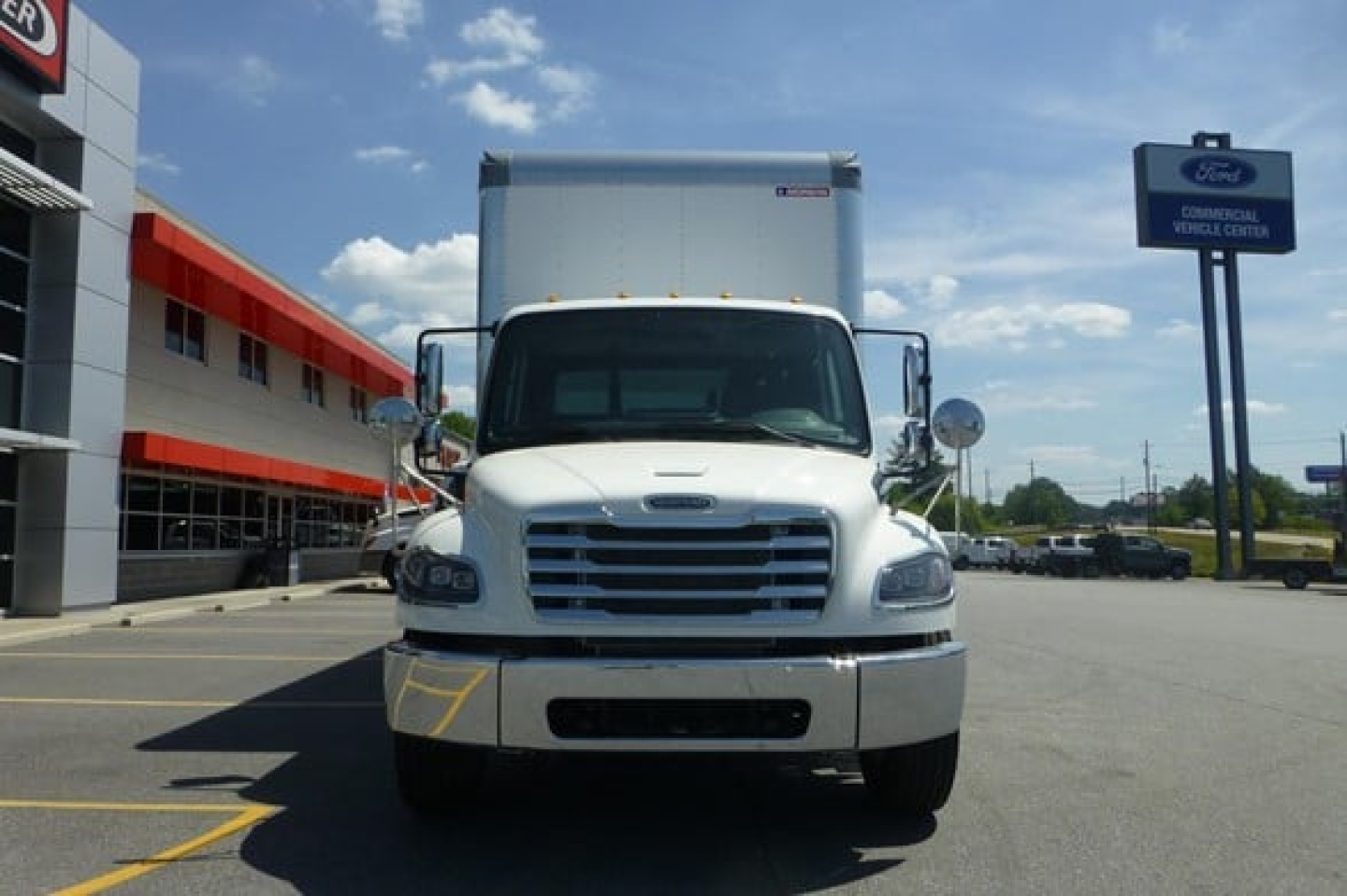 Freightliner M2 106 Plus box truck front view showing white cab, chrome grille, and cargo box, with dealership lot in background.