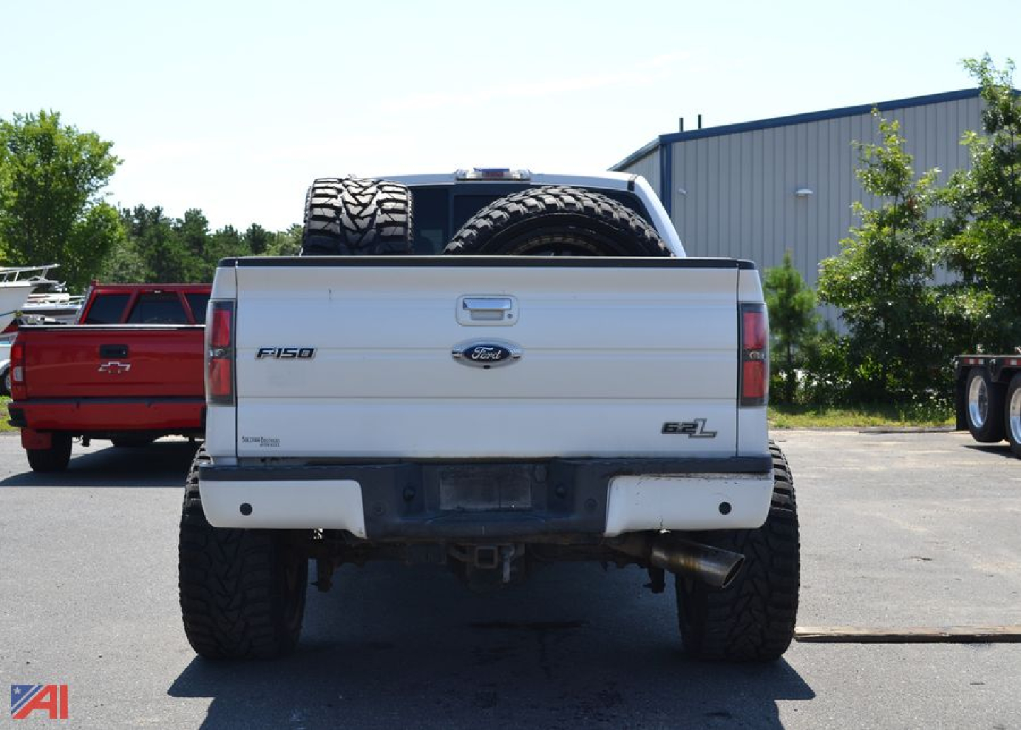 Rear view of a white 2011 Ford F-150 Crew Cab pickup truck showing spare tire mounted on bed and dual exhaust, with a red truck in background.