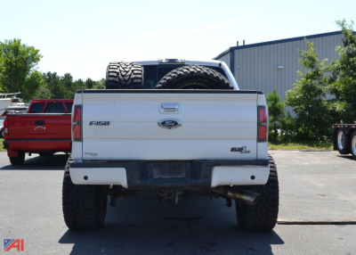 Rear view of a white 2011 Ford F-150 Crew Cab pickup truck showing spare tire mounted on bed and dual exhaust, with a red truck in background.