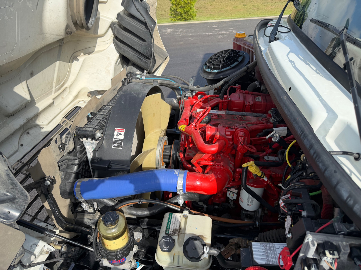 Engine bay of a Freightliner M2-106 showing red engine, blue coolant hose, yellow cap, black air intake, and various hoses and reservoirs.